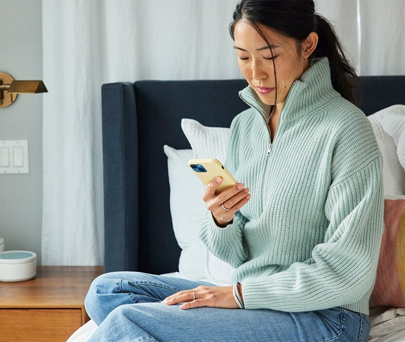 Une femme assise sur un lit, regarde son téléphone et réfléchit à la façon de créer un mot de passe mémorable.