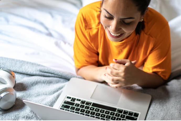 Jeune femme souriante dans son lit qui regarde son ordinateur portable.